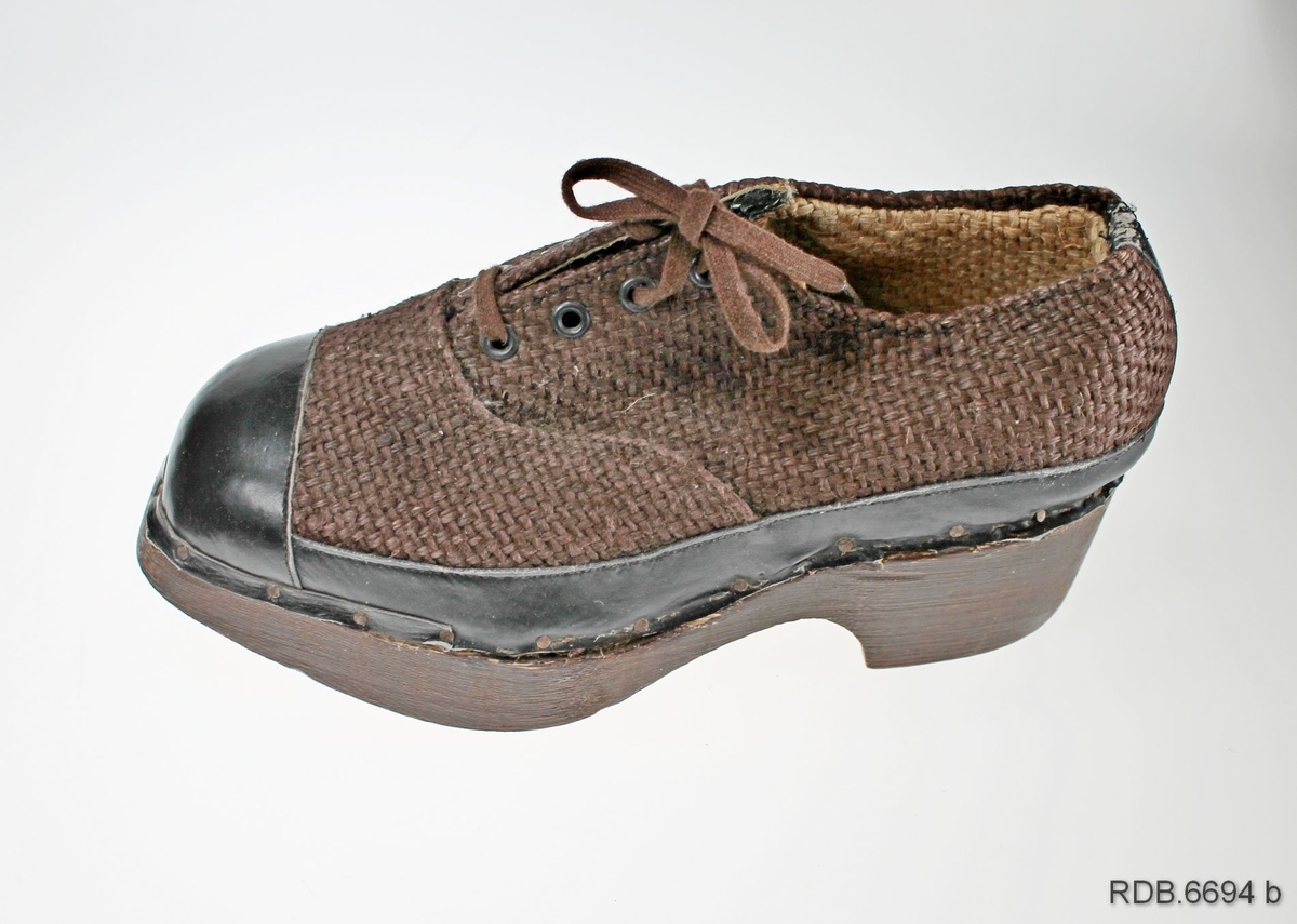Et par brune barnesko. Skoene er "krigsvare", fra 2. verdenskrig, med overlær av strie og tresåler (trebunner). Foran på tåa, på hælen og rundt kanten på skoen er påsydd en forsterkning av svart kunstlær som er spikret fast til trebunnen. Foran på skoen er det åpning med tunge under og 3 par hull med maljer, med brune skolisser er tredd gjennom. Under sålen er fastspikret 9 gummiknotter, tre av dem på hælen. Skoene er ikke brukt.