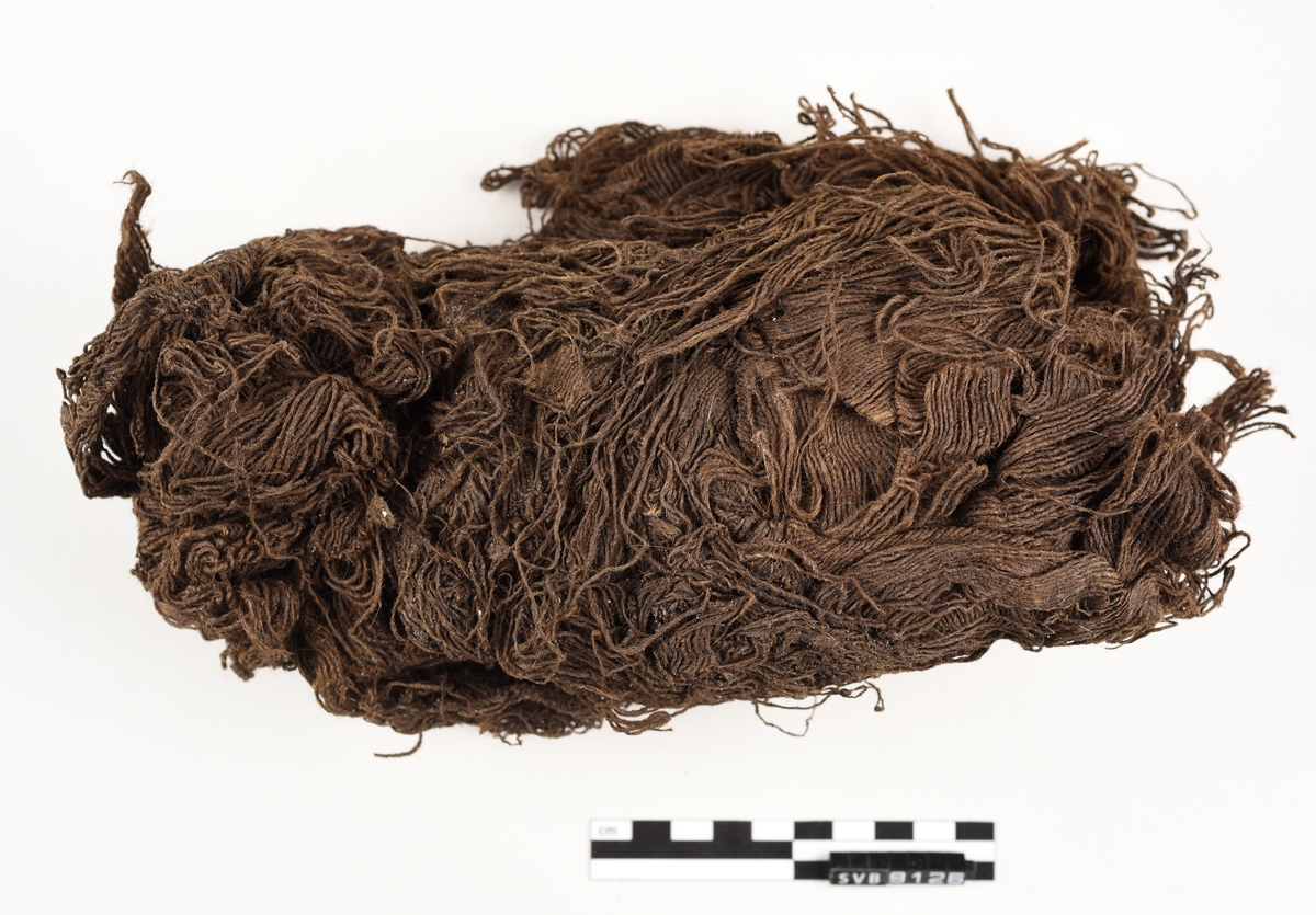 En bunt med tråd der fibrene er spunnet sammen. Det går en trepinne inn i gjenstanden. Gjenstanden er mørkebrun i farge. Gjenstanden er skjør.