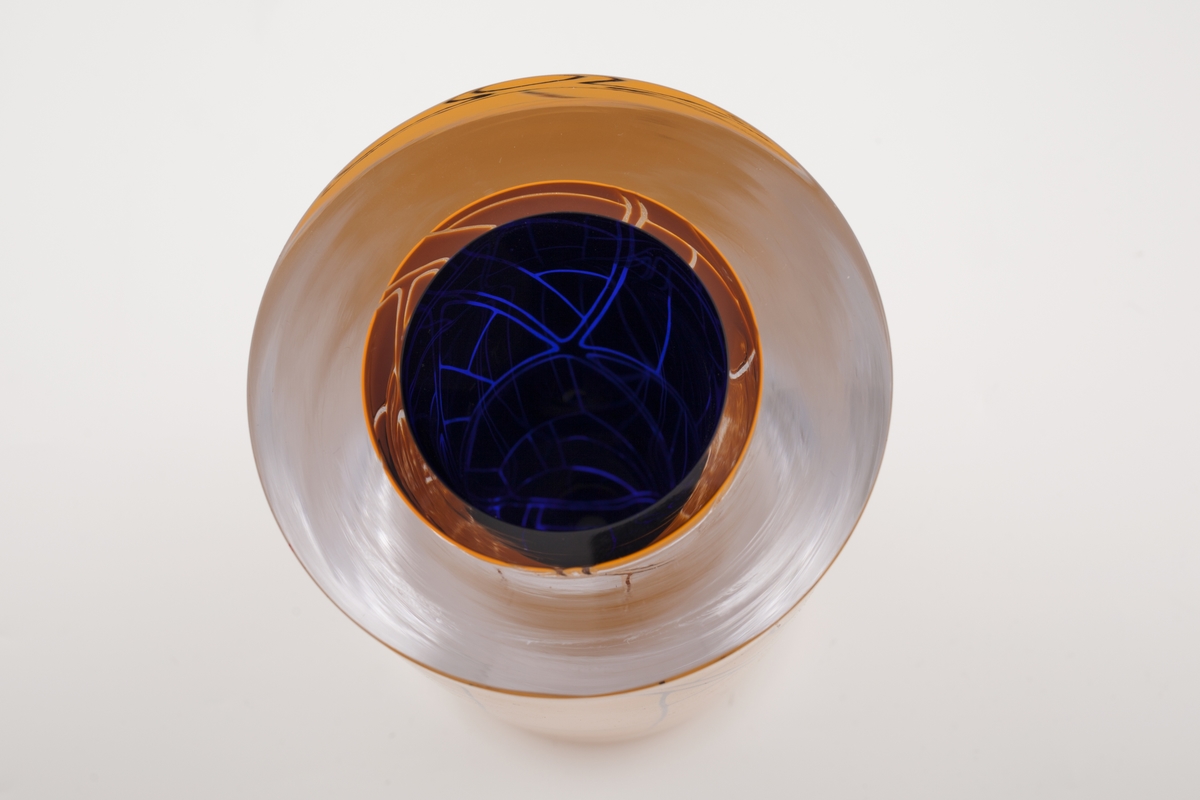 Massiv sylinderformet vase i underfangsglass, bestående sjikt i dyp blått, klart og orangefarget glass. Det orange sjiktet er delvis fjernet, slik at det blåfargen kommer til syne i form av geometriske linjeføringer. Planslipt topp og sirkulær åpning.
