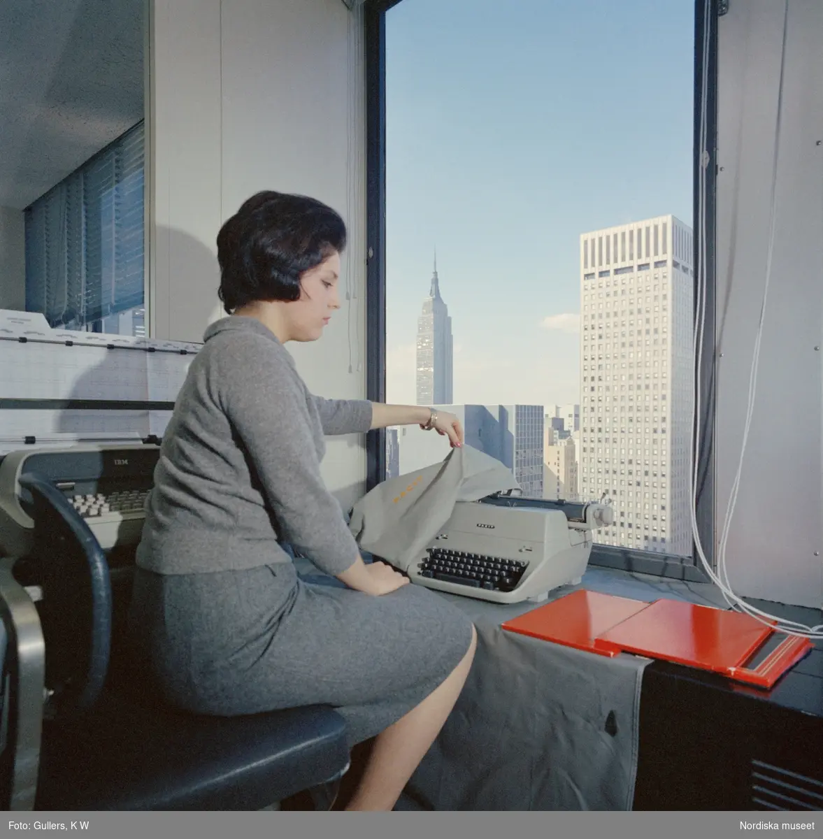 Facit-New York, 1960-tal. En kvinnlig modell avtäcker en Facit skrivmaskin sittande  invid ett fönster med utsikt över Empire state building.