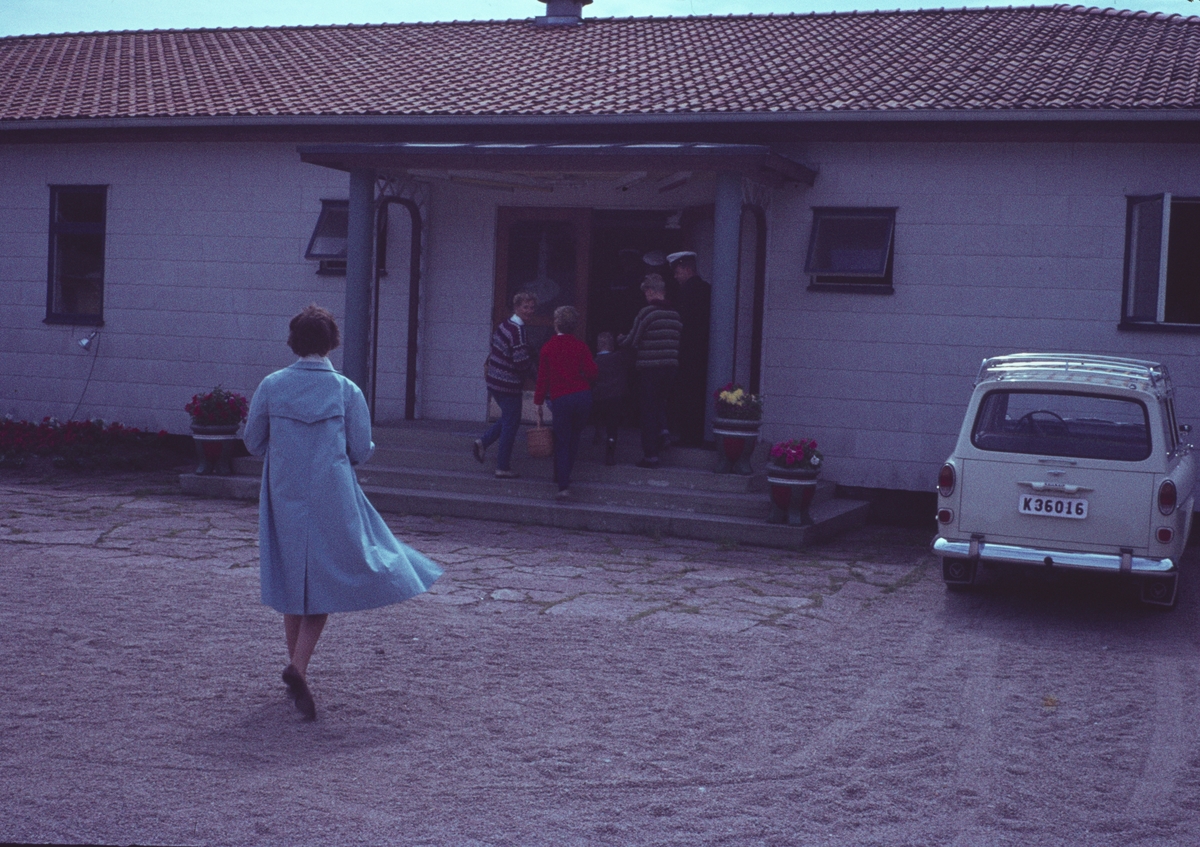 Bilden visar en kvinna i blå kappa som är på väg mot ett hus. Flera soldater och barn håller på att gå inomhus. På höger sidan parkera en bil.