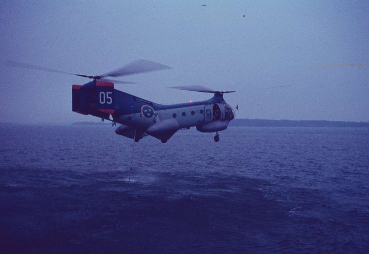 Bilden föreställer en så kallad flygande banan - en helikopter typ Vertol 44 som svävar knappt över vattenytan. Från bakdelen hänger en wire ner i vattnet. Förmodligen är det en hydrofon för ubåtsjakt.