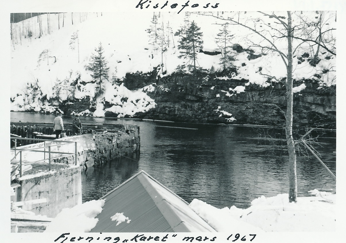 To personer står på muranlegg i Randselva på Kistefos Træsliberi. Taket til båthuset i forgrunnen.