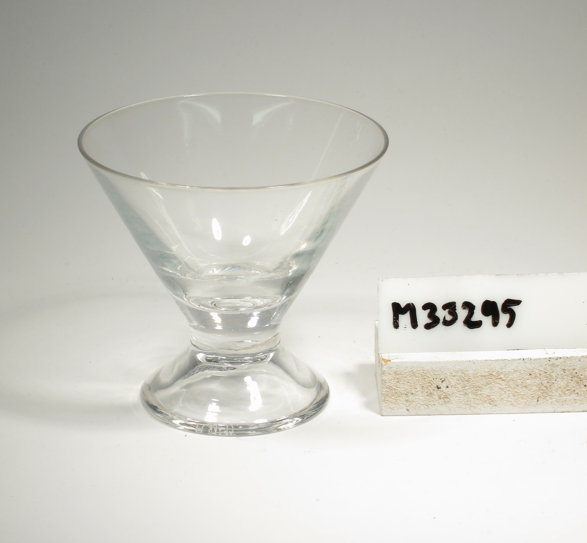 Likörglas med konisk kupa och påklippt ben. 
Etikett: Rektangulär med rundade hörn, silverfärgad med blå text: "Artium"