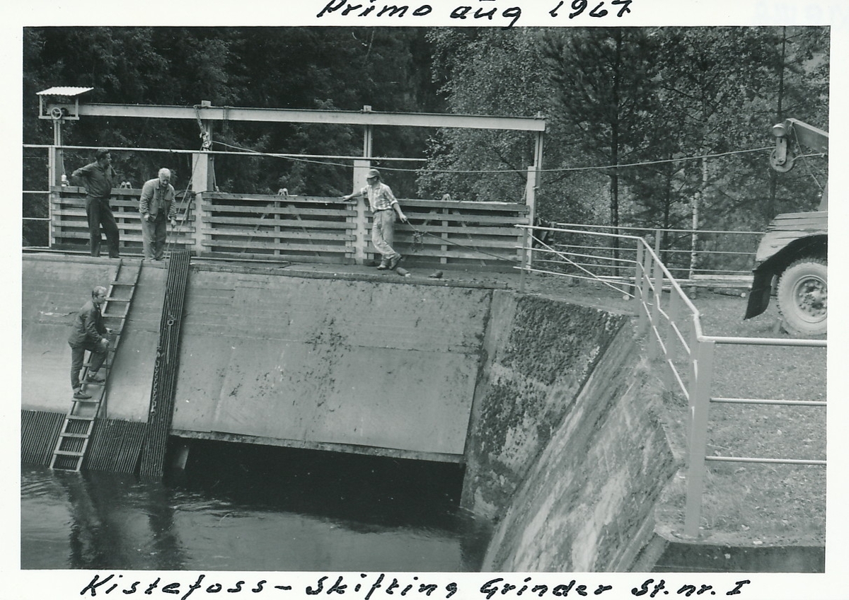 Demningen på Kistefoss under skifting av grinder i dammen. En mann står på stige ned i vannet, mens tre menn er oppe på demningen. Lastebil med kran på lasteplanet til høyre.