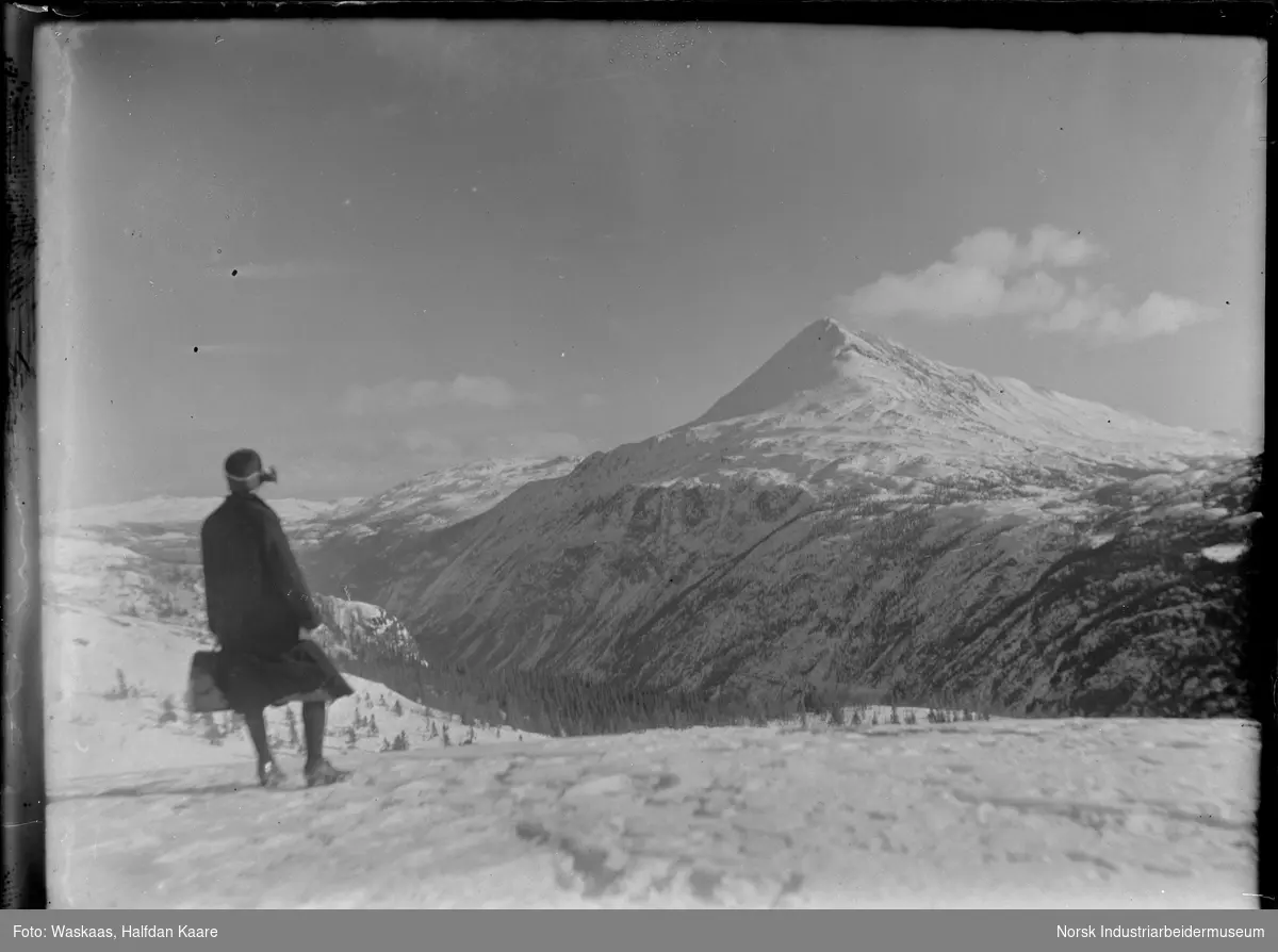 Kvinne i kjole og hatt stående på Pignatten (?) med utsikt til snødekket Gaustatoppen på vestsiden. Utsikt innover Gausdalen.