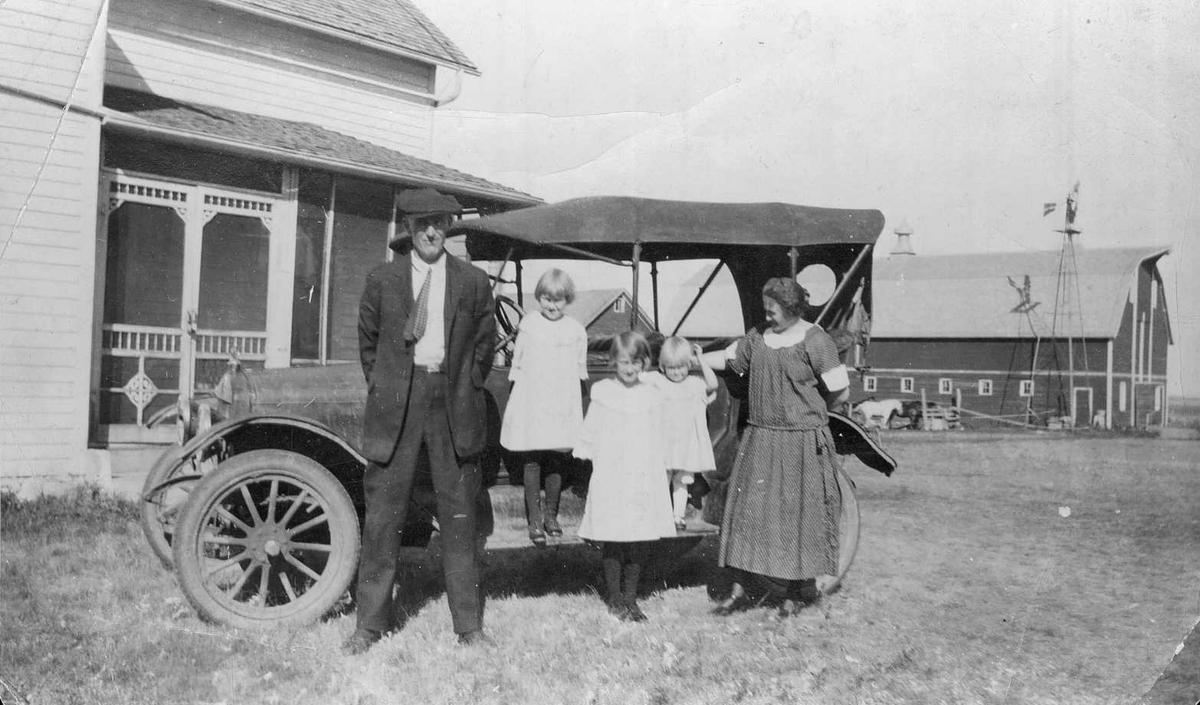 Amerikabilde, familie framfor bil, i bakgrunnen låve med vindmølle