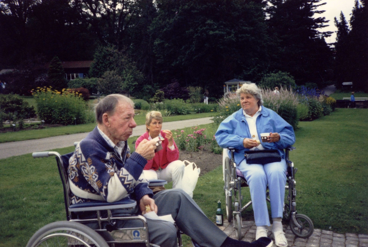 Brattåsgårdens ålderdomshem är på utflykt till Botaniska trädgården, 1980-tal. Från vänster: okänd man i rullstol, vårdbiträde Britt Fredriksson samt Elsa Kristiansson (1925 - 1994) i rullstol.