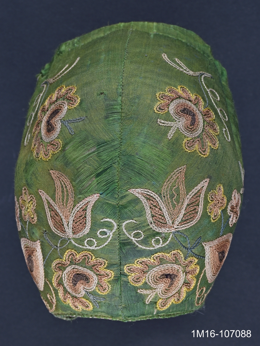 Bindmössa av grönt siden med gulbrunt tambursömsbroderi i bruna nyanser. Bindmössan kopierades till Valledräkten under 1960-talet av ett dräktråd. Har tillhört Carolina Johansson (1835-1917).