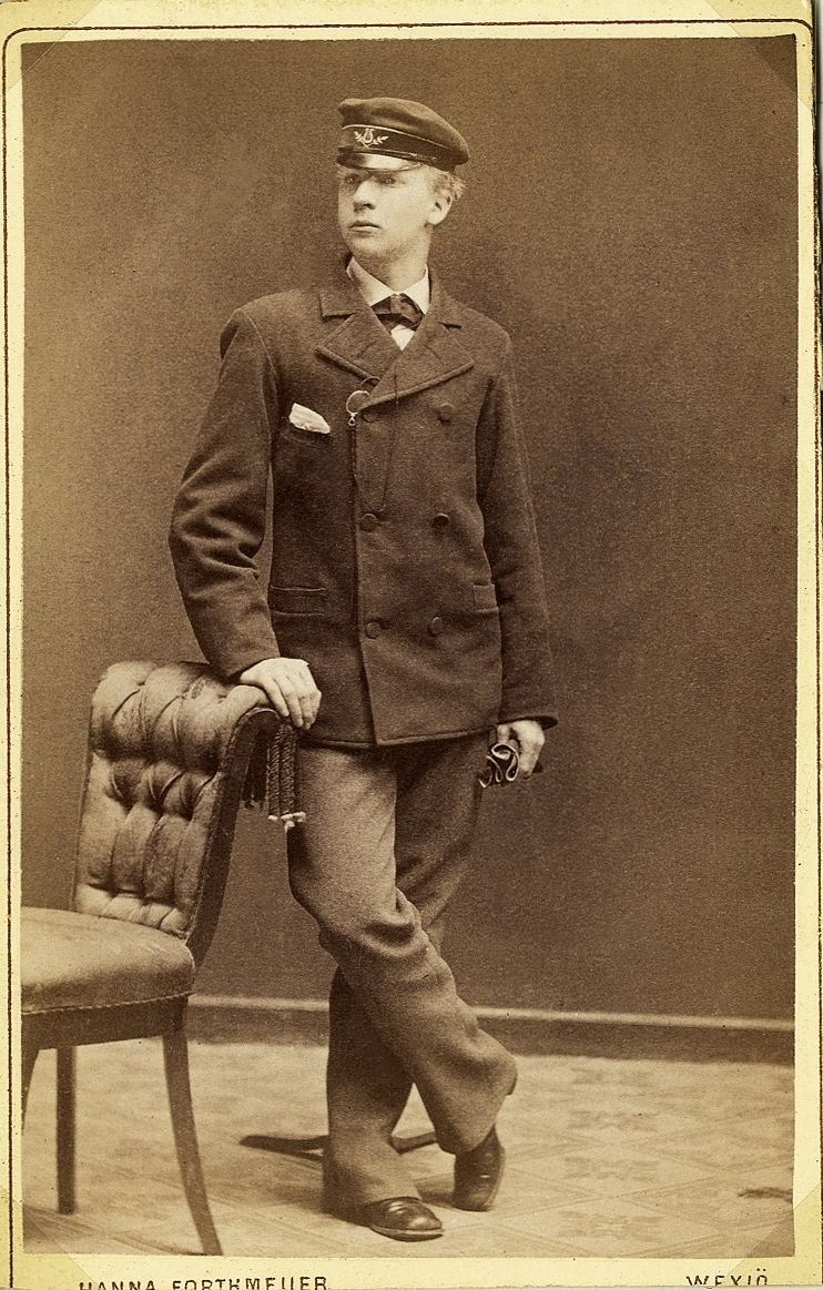 Foto av en ung man i kavajkostym med fluga och skolmössa. Vid kavajslaget skymtar en pincené med snodd. 
Han lutar ena handen mot ryggstödet på en stoppad stol. 
Helfigur, halvprofil. Ateljéfoto.