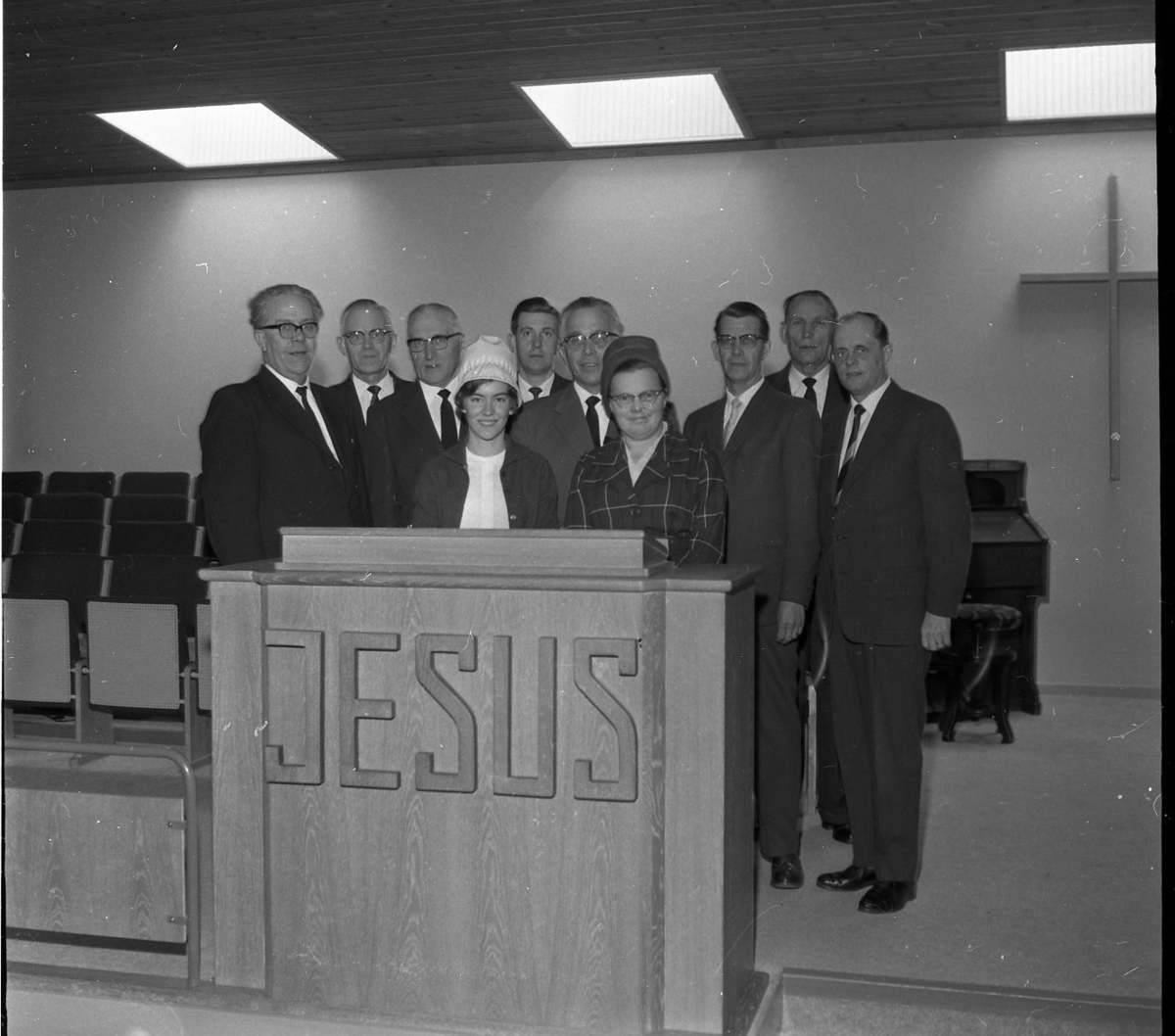 Åtta kostymklädda män och två kvinnor med hatt står bakom talarstolen i Filadelfiakyrkan. På densamma, mot salen, står texten "JESUS". Mannen som står mellan kvinnorna är pastor Robert Svensson.