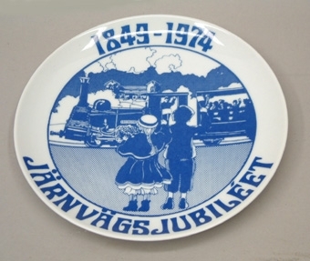 Vit porslinstallrik med blått tryck. Text: "1849-1974 Järnvägsjubileét". I mitten en illustration  tecknad av Åke Arenhill, som visar två barn i historiska kläder som tittar på loket Frykstad som drar en personvagn.