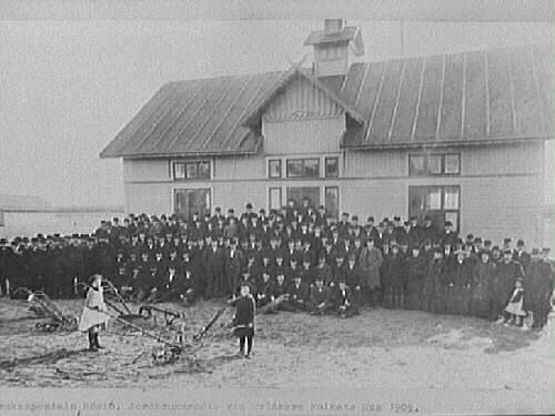 Text till bilden: "Småbruksaposteln Rösiö. Jordbrukarmöte vid Tvååkers Folkets hus 1905." En mycket stor samling män och några kvinnor står uppställda framför huset. I förgrunden står ett antal jordbruksmaskiner och två små barn.