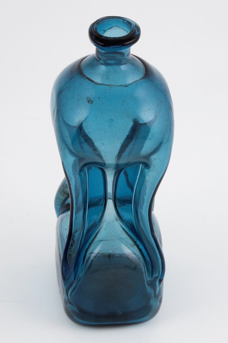 Klukkflaske i gjennomskinnelig mørkeblått glass. Timeglass-lignende utforming, hvor den tilnærmet kvadratiske over- og underdelen er forbundet med et tynt rør. Lav hals med glatt kantring, samt puntemerke på undersiden.