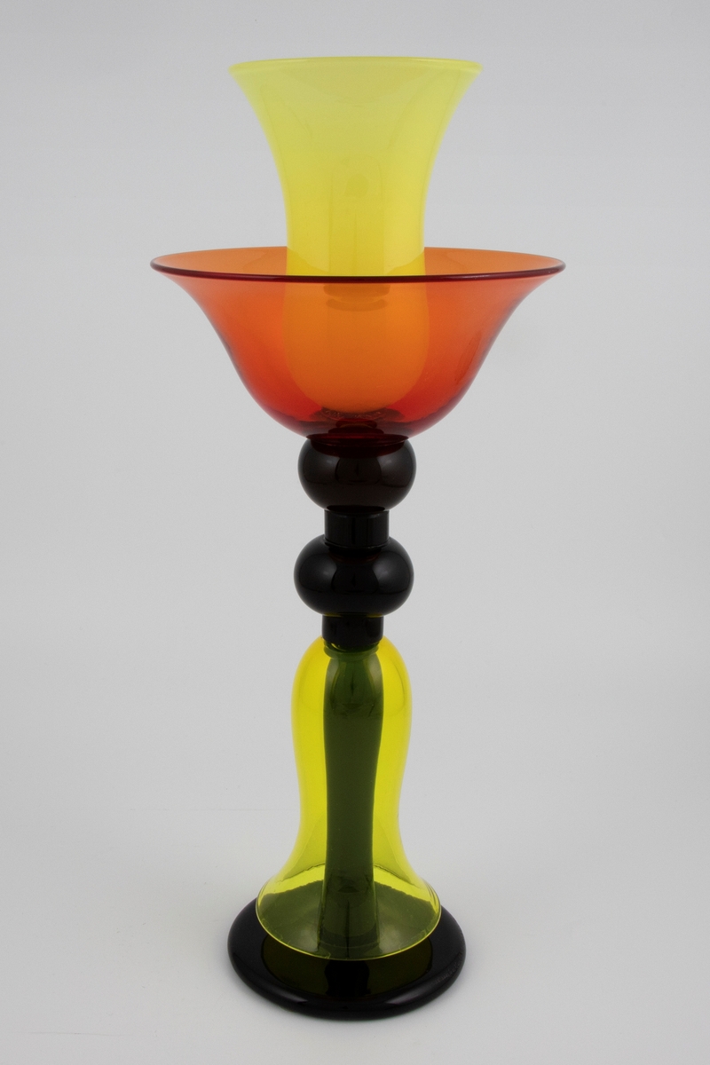 Høyreist vase i farget glass. Skålformet kupa i orange, hvorfra det reiser seg en mindre balusterformet vase i halvgjennomskinnelig lysegult glass. Flerleddet kuleformet stett og sirkulær fotplate i opakt sort glass. Nedre del av stetten er dekket av en balusterformet klokke i gulfarget glass.