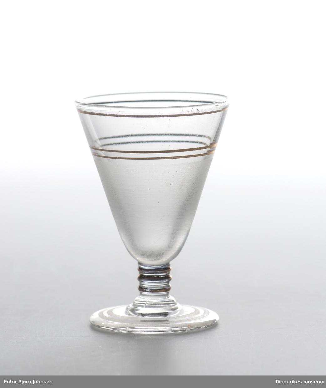 Fire akevittglass dekorert med tre gullstriper øverst på glasset tre tykkere gullstriper på stetten. Nederste del av glasset er etset og foten har to tynne etsete striper.