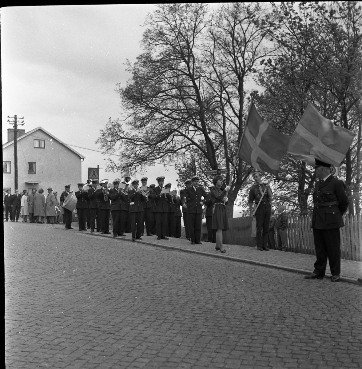 En musikkår med fanor i täten står uppställd vid Södra tullen i riktning mot staden. Polis Holger Isacsson står till höger. Fastigheten Jönköpingsvägen 2 syns i bakgrund, skyltar med "Rum" att hyra.