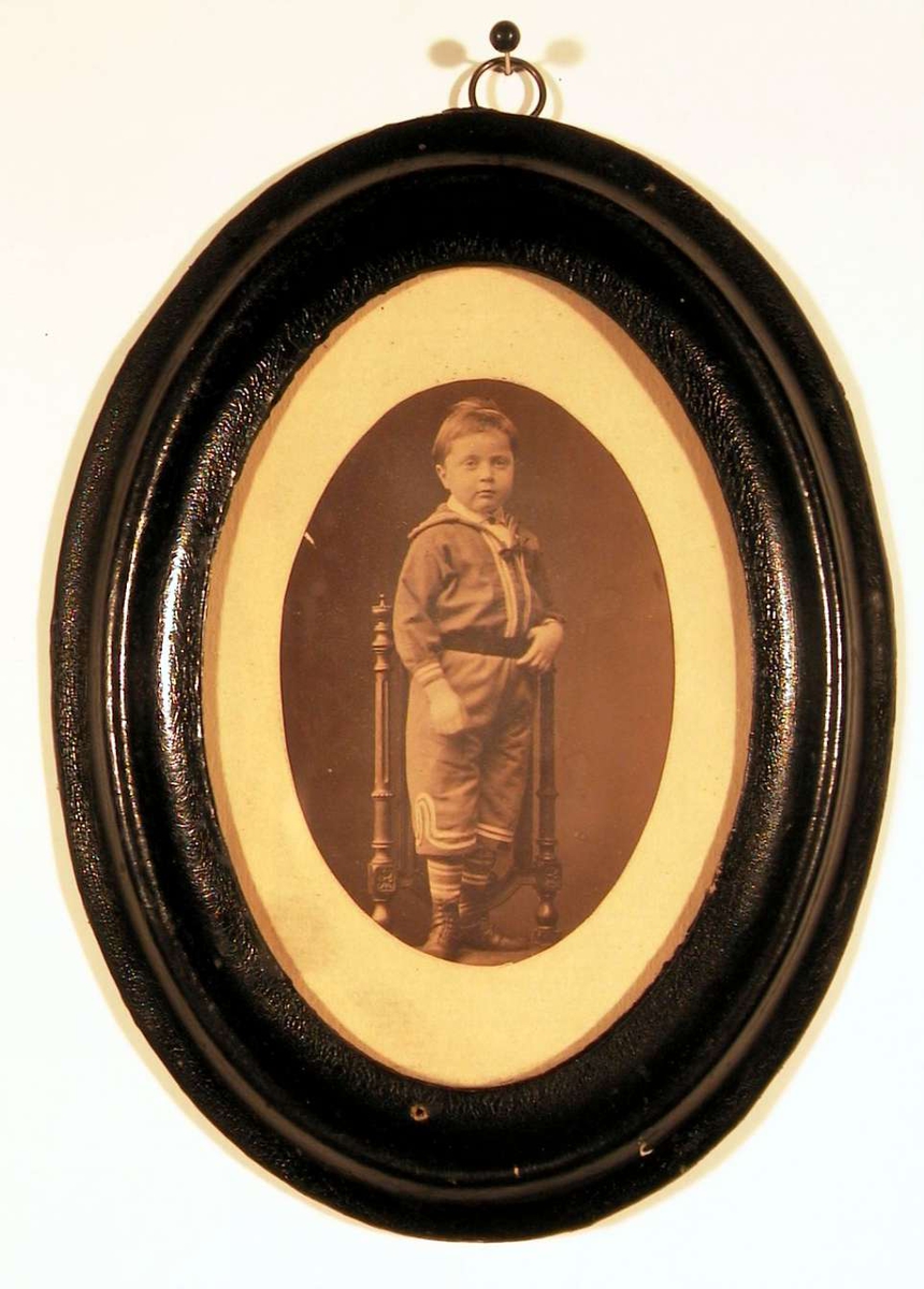 Fotografi av gutt stående på stolsete, kledd i matros drakt med bredt mørkt belte om livet, stripete strømper og støvler. Han ser direkte på betrakteren.