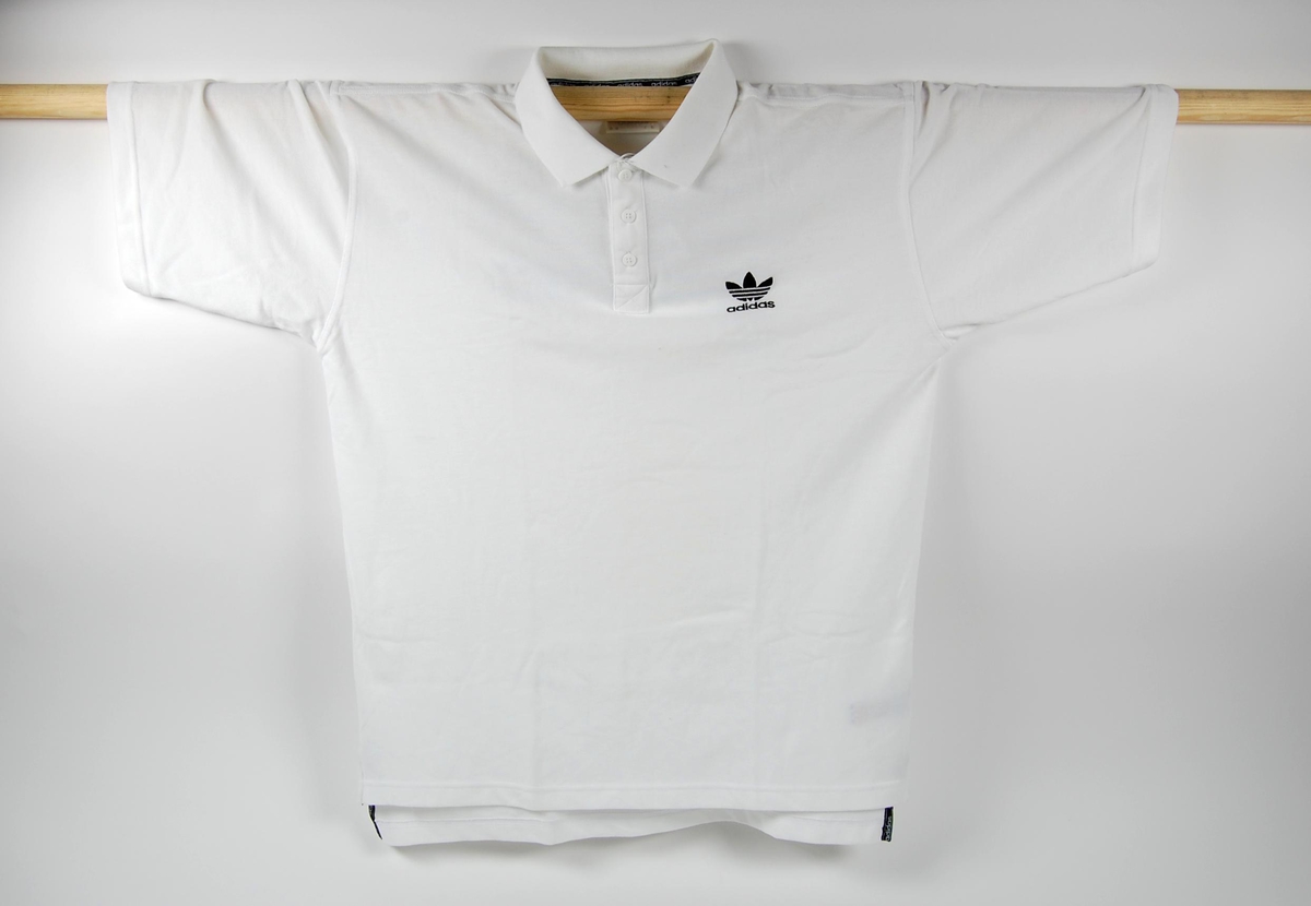 Hvit t-sjorte med blå innskrift på ryggen. Sort logo for Adidas på brystet. T-skjorta har knepping ved brystet.