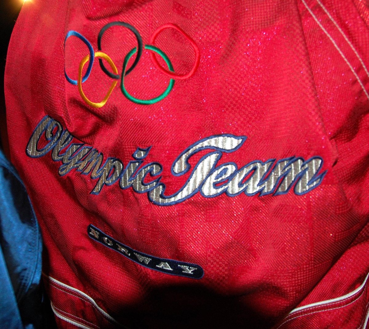 Rød jakke med hvite og blå striper. Jakken har tre lommer. De olympiske ringene i farger brodert på ryggen.