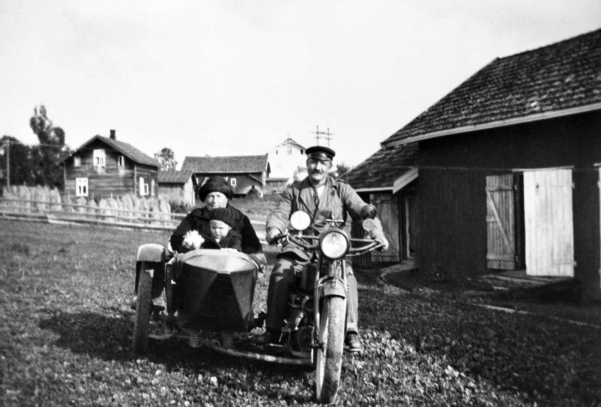 Motorsykkel med sidevogn. Mann på sykkelen med kvinne og barn i sidevognen. Oppstilt ved et uthus, i bakgrunnen et hus og korn på staur. Ukjent sted