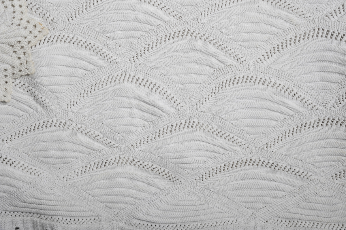 Strikket sengeteppe med mønstereffekt i vifteform. Rett og vrangmasker utgjør mønster. Rundt teppet er det påsydd en strikket kant.