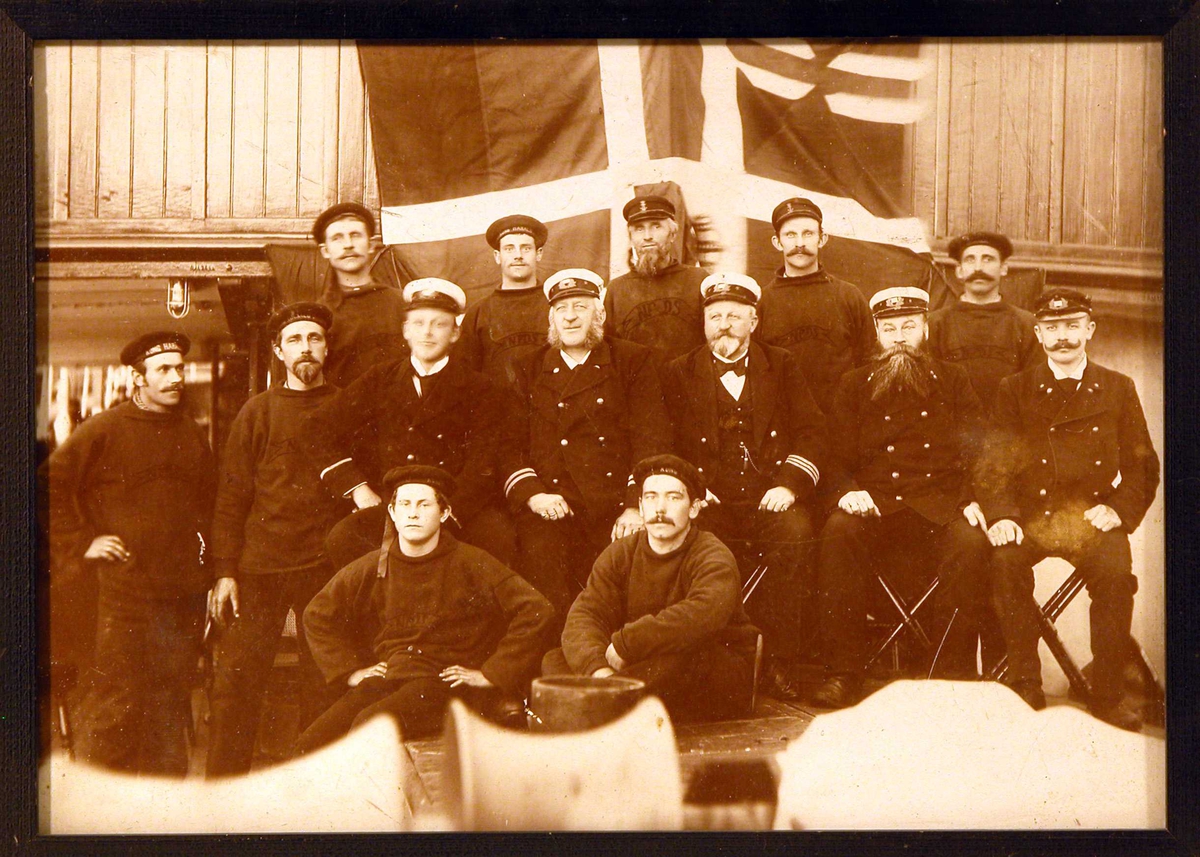 Gruppebilde av mannskap på D/S "Kong Harald" (NFDS) poserende på skipsdekk foran det svensk-norske unionsflagget ("sildesalaten"). 
Bildet er tatt rundt århundreskiftet 1900 +/- 5 år.