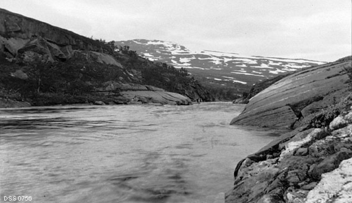 Utløpet av Store Kalvatnet, sett i medstrøms retning.  Bildet viser hvordan vatnet renner ut av Kalvatnet mellom to bergflater, dels blankskurte, men med en del bjørkevegetasjon på venstre side.  I bakgrunnen en fjellrygg med snøflekker. 