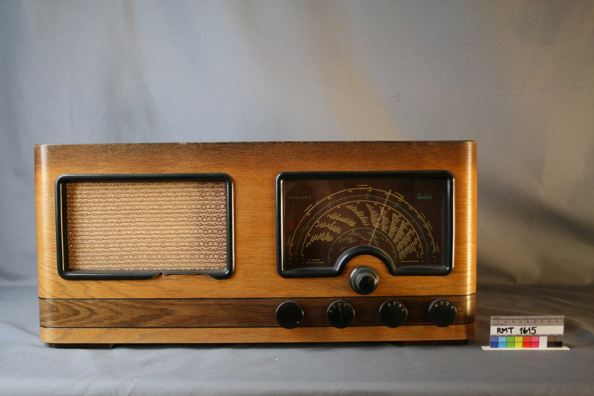 Rektangulær brun radio. Forside til høyre: skala med fem funksjons- og søkeknapper. Til venstre høyttaler med trekk av tekstil. Bakside: bakvegg mangler på venstre side. Forskjellige uttak.