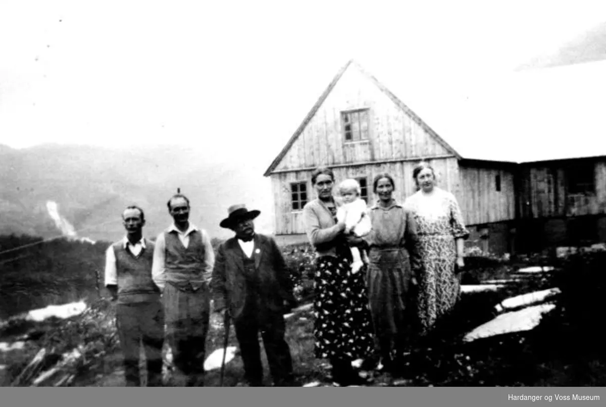 seks personar framfor bustadhus, Tvinnefossen i bakgrunnen