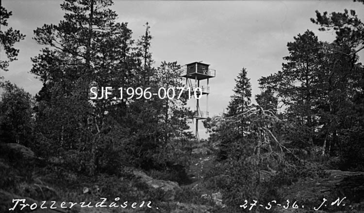 Skogbrannvaktstasjonen på Trollerudåsen i Spydeberg.  Brannvakttårnet på dette stedet ble reist i 1935, som en erstatning for eldre brannvaktposter på Kjerringa, Spydeberg varde og Bjørnåsen.  Trollerudås-tårnet er i hovedsak bygd av stål og betong, værbestandige materialer som skulle gi tårnet lang levetid.  Konstruksjonen består av fire loddrette betongsøyler med tre horisontale avsatser mellom.  Samtlige avsatser har stålrekkverk. På den øverste avsatsen er det bygd ei lita utkikkehytte av tre.  Atkomsten til hytta skjer ved hjelp av stiger som er ført gjennom åpninger i de ulike avsatsdekkene.  Dette fotografiet er tatt på avstand og viser hvordan tårnet er plassert på et berg der det vokser litt forkrøplet furuskog i revnene. 