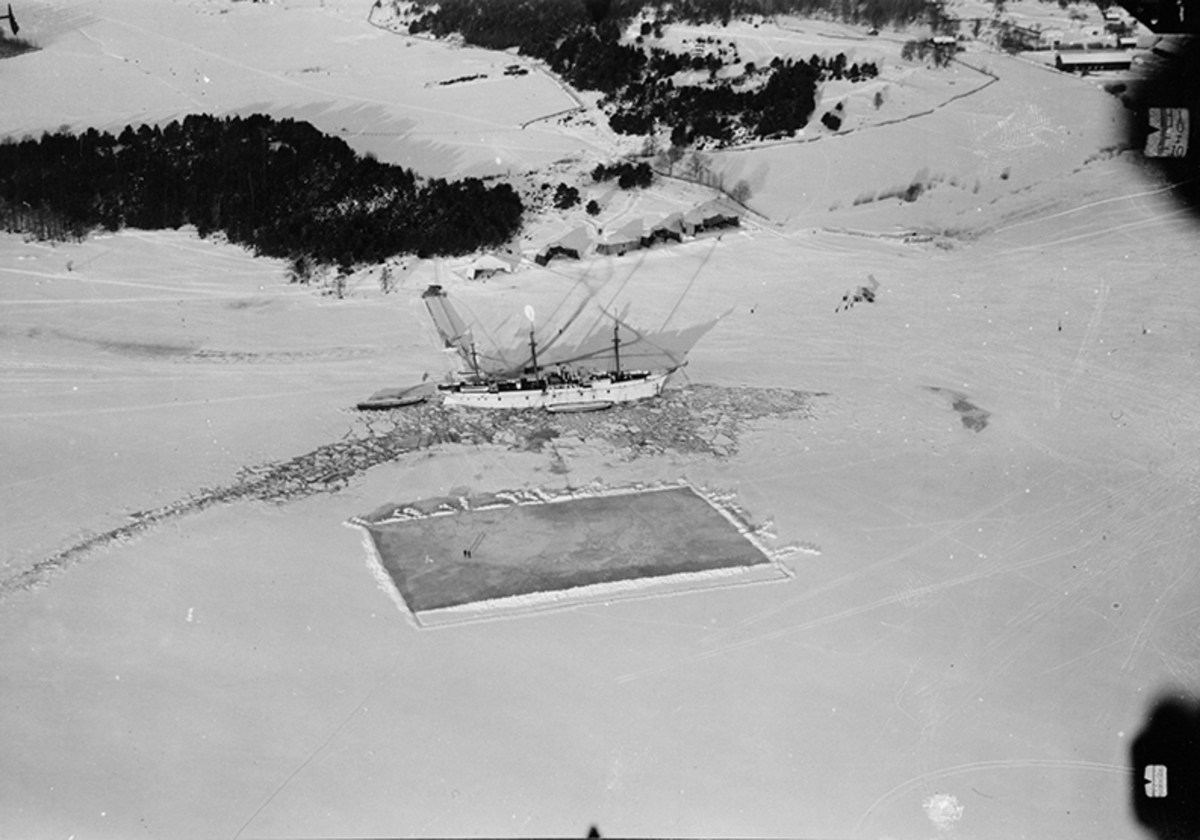 Logementsfartyget Freja förankrat i isen utanför bryggan vid Hägernäs flygstation. Uppskottad bandyplan på isen. Tälthangarer på strandkanten.