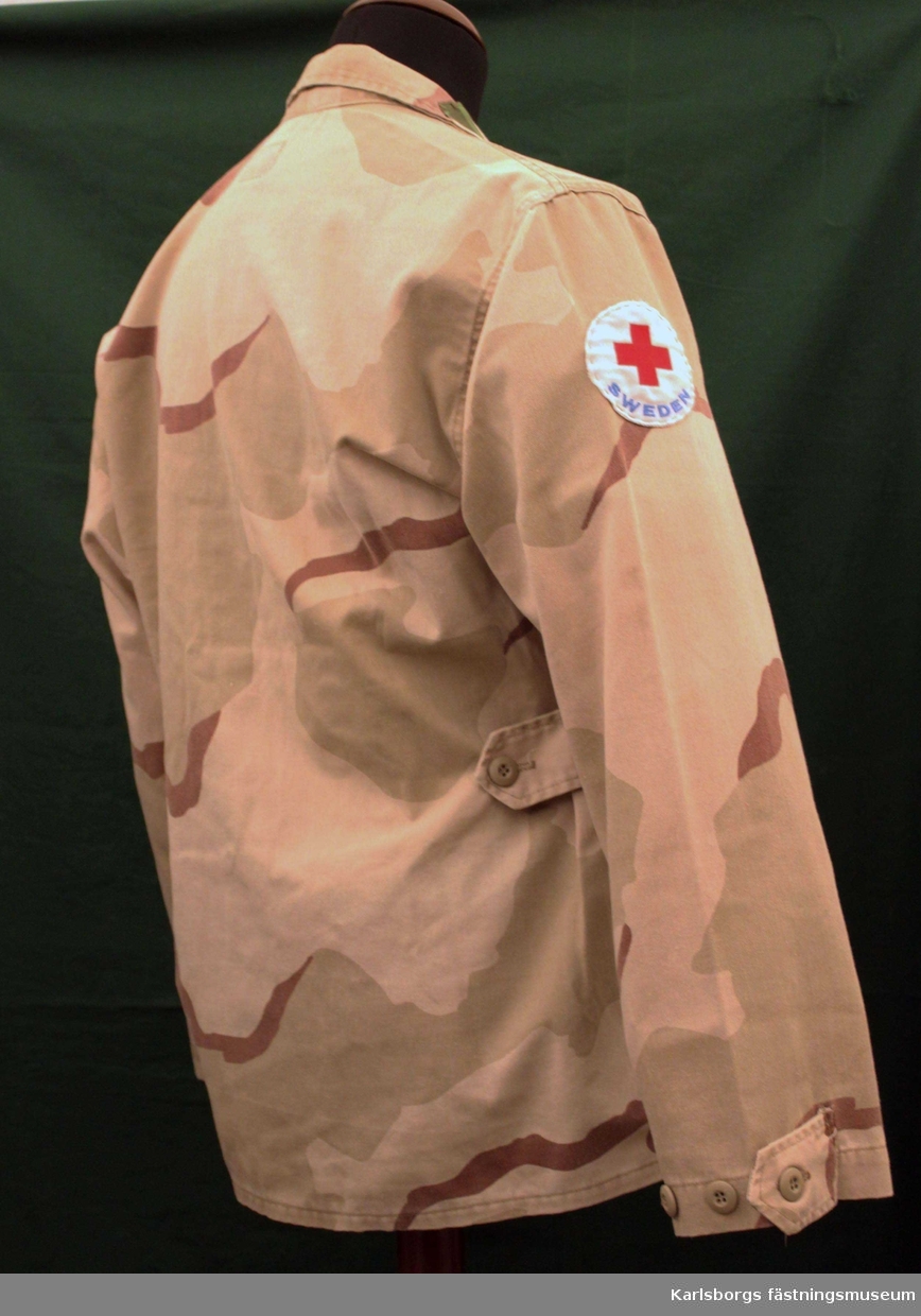 Kamouflagejacka,enkelknäppt med doldknäppning.och skjortkrage. Utanpåsydda fickor med ficklock, Röda korsemblem på höger ärm samt märke med tre kronor på vänster ärm.