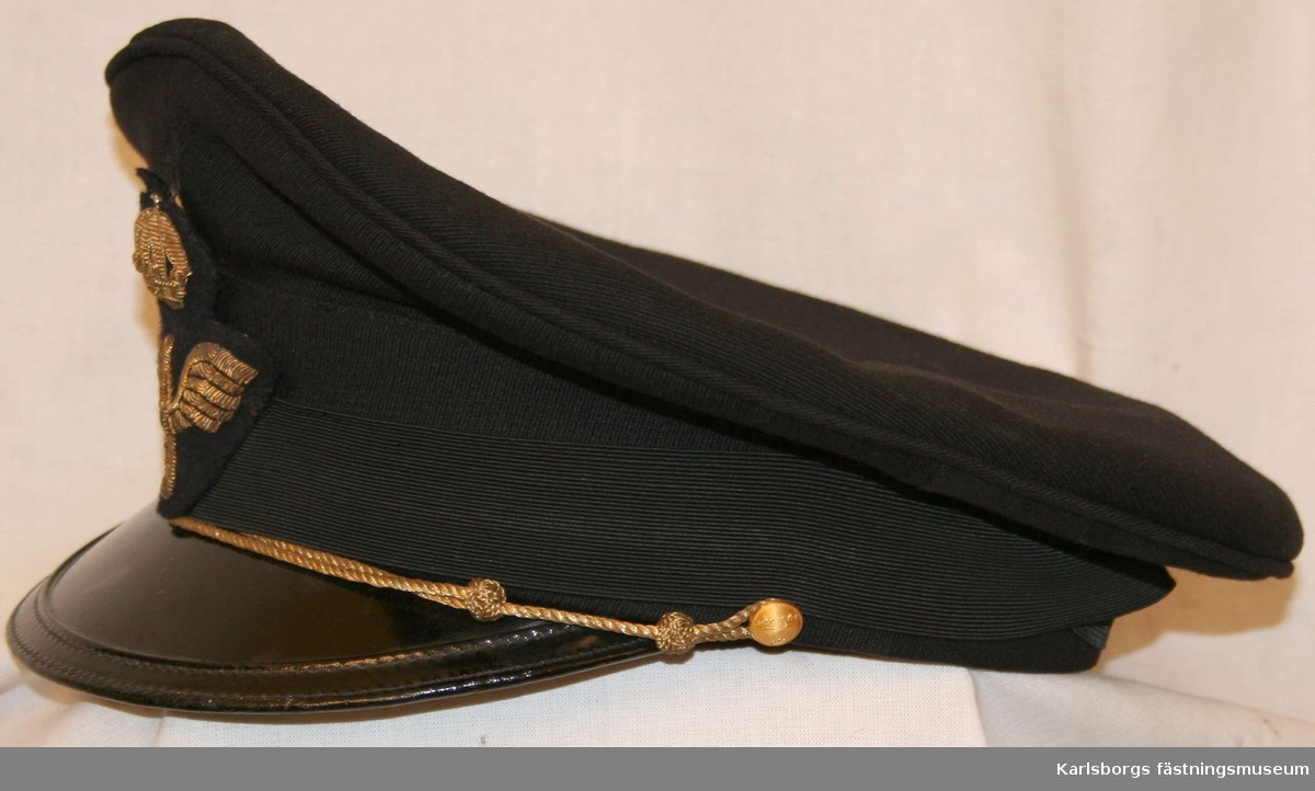 Storlek: 56
Skärmmössa m/1930 av mörkblått tyg mössband av svart räfflat silke. Svart lackerad skärm, försedd med hakrem av guldsnodd fäst vid två 14 mm uniformsknappar samt flygemblem.