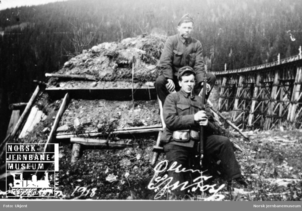 To militære foran ei torvhytte med Drøia bru i bakgrunnen