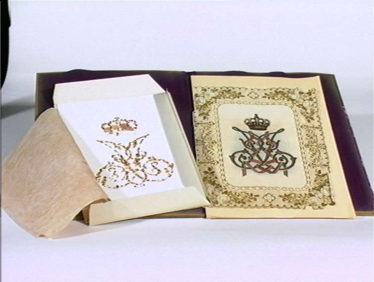 Mappe av tre. Utskåret gjennombrutt rankeornamentikk, kronet dobbeltmonogram, dronning Josephine, og årstall 1824 - 1874. Innvendig foret med fiolett silke.