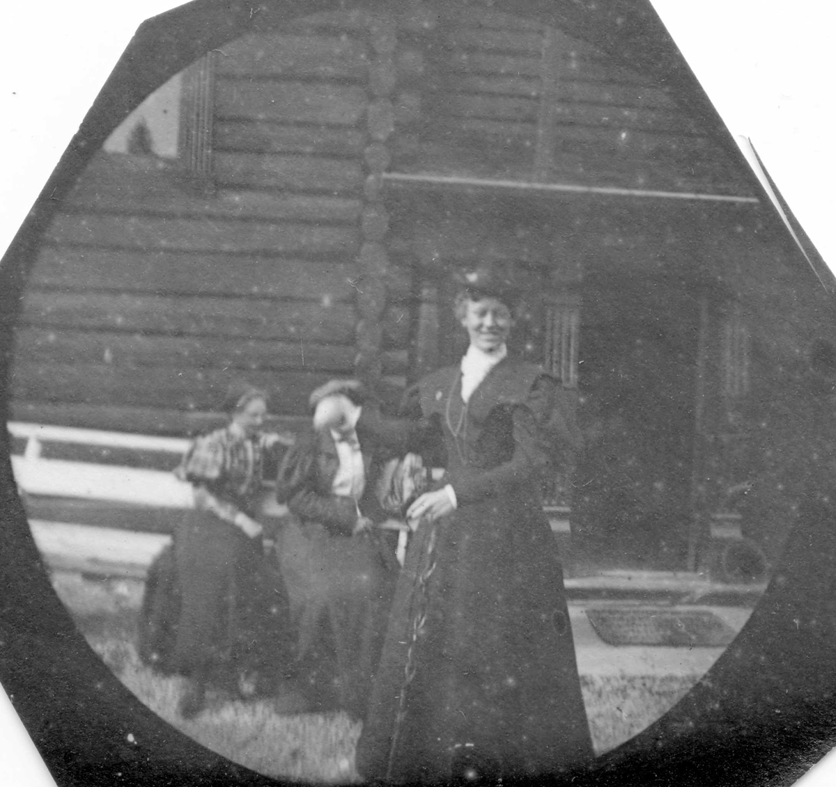 Golå, Sør-Fron, Oppland. Frk Alnæs står foran tømmerhus. To damer sitter på krakk.