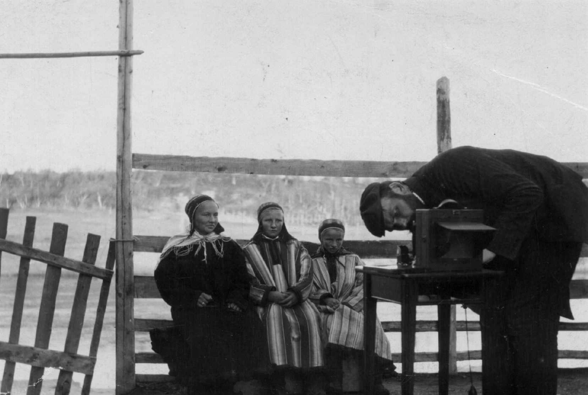 Språkforsker Konrad Nielsen (1875-1953) ved fotoapparatet, tre samiske kvinner på en benk i bakgrunnen. Karasjok. Forsker fotograferer.