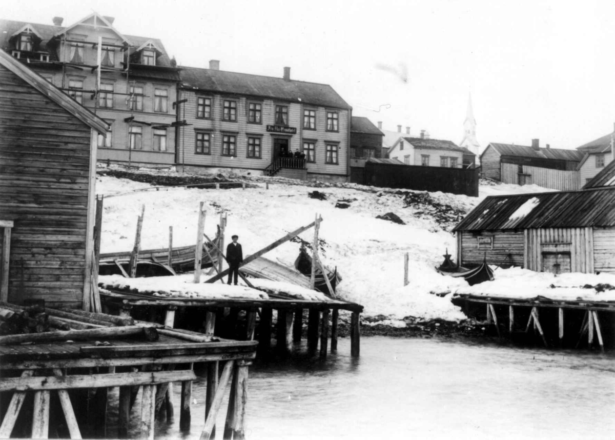 Vadsø, Finnmark. Havneparti med bebyggelse og båter trukket opp på land. Fru Aas hotel i husrekken og kirkebygning i bakgrunnen.