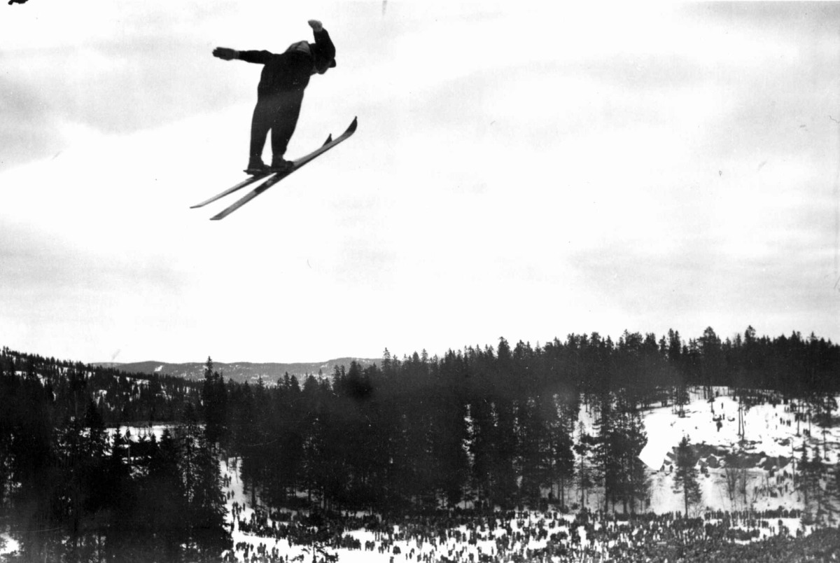 Ski. Hopprenn. Holmenkollen, Oslo 1934. Hopper i svevet. Publikum og Nordmarka ses i bakgrunnen.