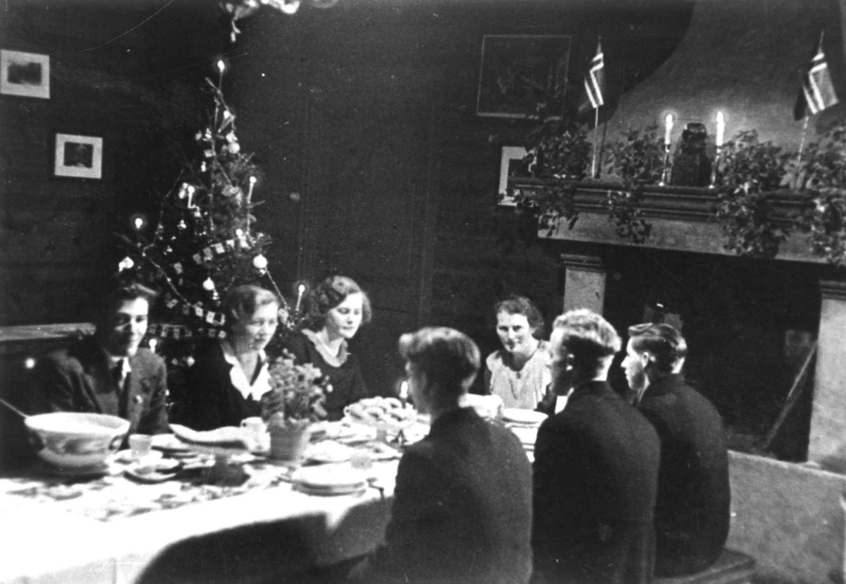 medlemmer av Anna Grostøls familie sitter rundt et dekket spisebord foran peisen. Et juletre i bakgrunnen. Vest-Agder 1941-42.