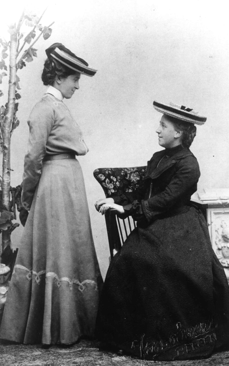 Kvinnedrakt, antatt 1900-årene. 2 kvinner poserer i fotoatelier, den ene sittende.
Fra familiealbum uten videre opplysninger.