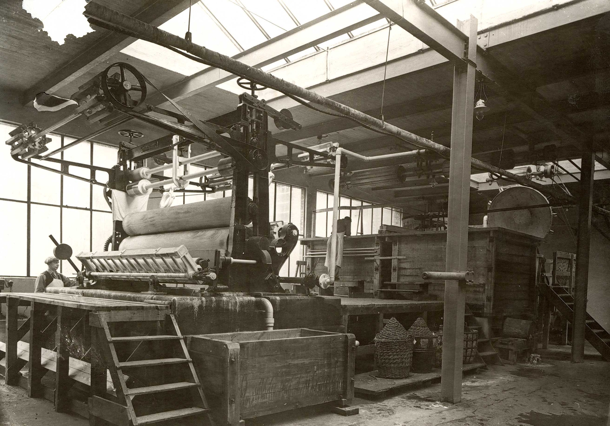 Produksjonslokale ved A/S Joh. Petersens Linvarefabrik. Mann ved maskin med store valser. Trekasser i bakgrunnen. Blekingen.