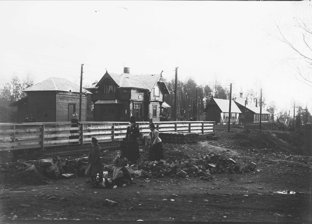 Ventende togpassasjerer foran toglinje med stasjonen i bakgrunnen. Sannsynligvis Slemdal stasjon, Holmenkollbanen.