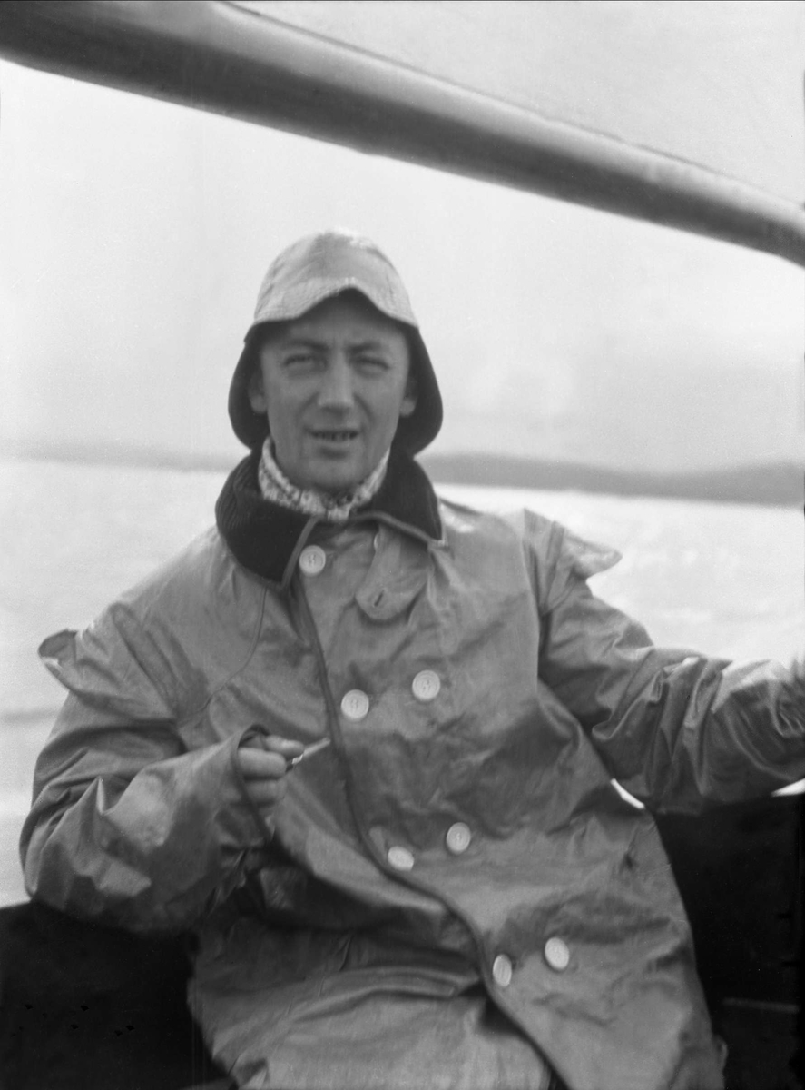 En mann med sydvest og pipe i hånden sitter i en seilbåt. Robsahm og Lund.