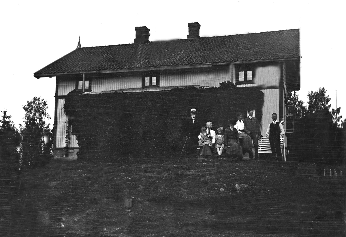 En gruppe mennesker, voksne og barn står oppstilt foran et trehus. Robsahm og Lund.