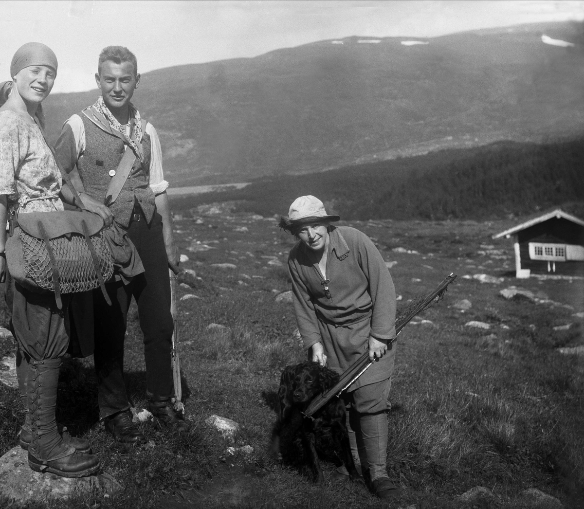 Gerd og Åge med fiskeutstyr står sammen med tante ant. Sussa som står med en hund og fiskestang på fjellet, en hytte i bakgrunn. Fotografert 13. mars 1926.