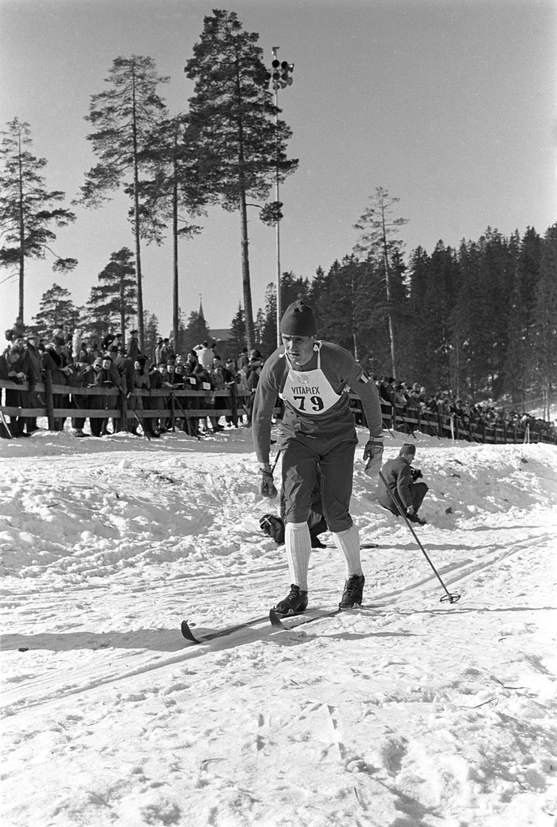 Skiløper med startnummer 79 i Holmenkollen. Publikum langs løypa.  Pressefotografer på sidelinjen. Holmenkollrennene 1963.