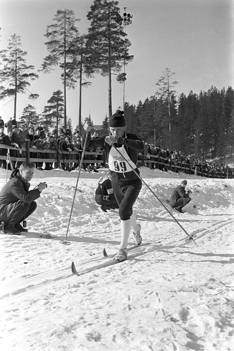 Skiløper med startnummer 99 i Holmenkollen. Publikum langs løypa.  Pressefotografer på sidelinjen. Holmenkollrennene 1963.