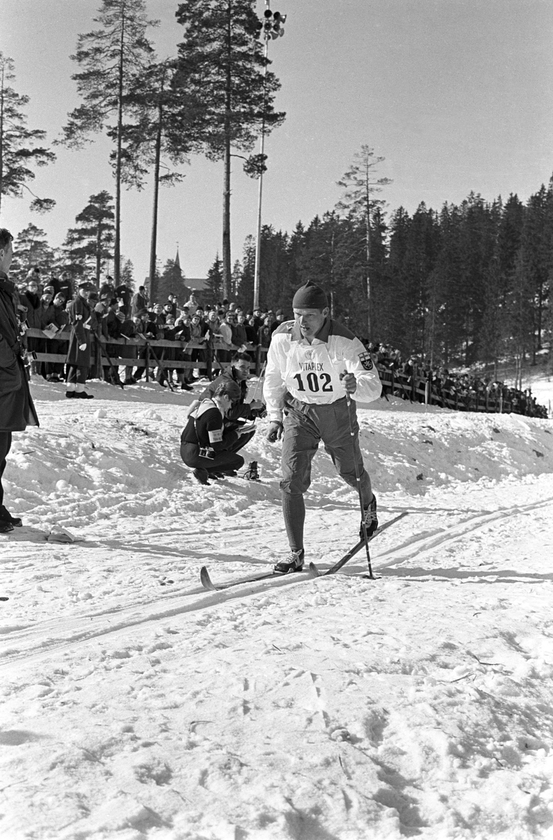 Skiløper med startnummer 102 i Holmenkollen. Publikum langs løypa.  Pressefotografer på sidelinjen. Holmenkollrennene 1963.
